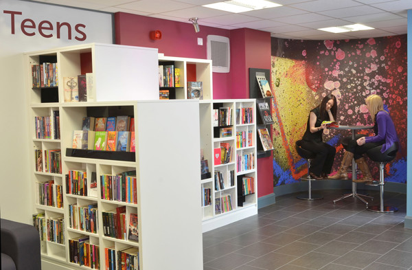 Two young people engaged in conversation at a high table beside colourful browsing shelves in the young readers' browsing space at Gateshead Central Library