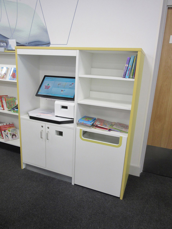 Self-service kiosk with a touchscreen and book return slot beside open shelving at Beeston Library