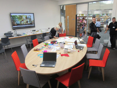 Oval wooden table surrounded by grey and red chairs, with laptops and craft materials for a makerspace activity at Beeston Library