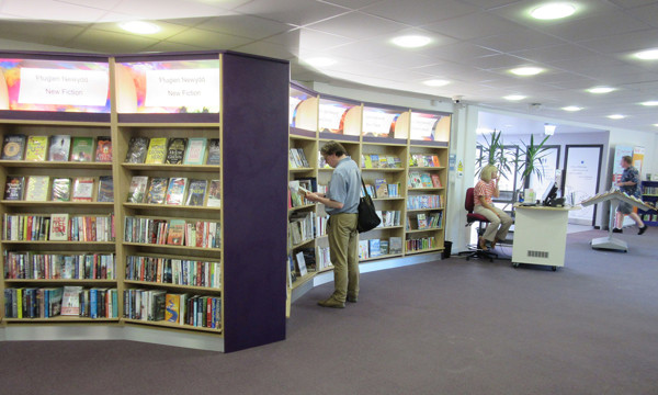 Curved shelving displaying new fiction titles with a patron browsing at Wrexham Library