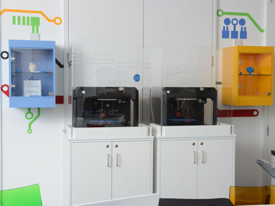 Two 3D printers housed in transparent enclosures beside a colourful wall display at Stafford Innovation Suite