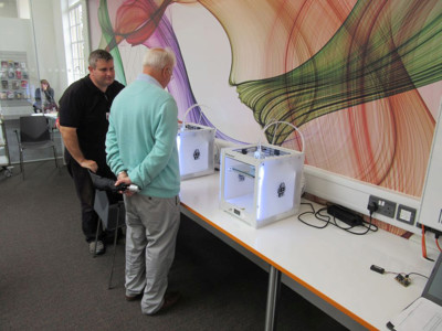 Two individuals observing 3D printers on a long table with colourful wall graphics at Beeston Library