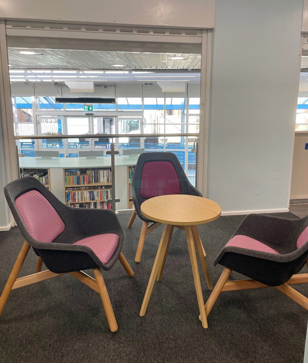 Two upholstered lounge chairs with pink and grey fabric beside a wooden side table in a quiet reading space at Birtley Library