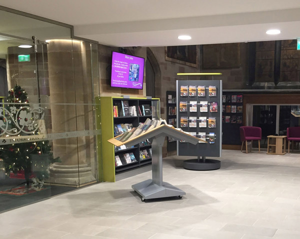 Leaflet display stand with angled shelves and browsing shelves beside purple lounge chairs at Lichfield Library