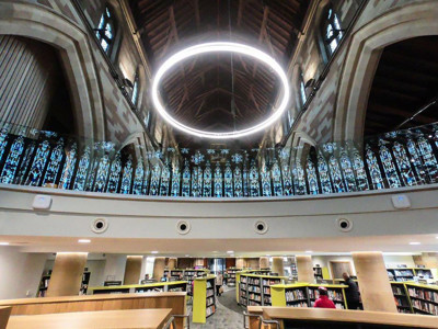 Illuminated circular light fixture above the open-plan library floor with browsing shelves at Lichfield Library