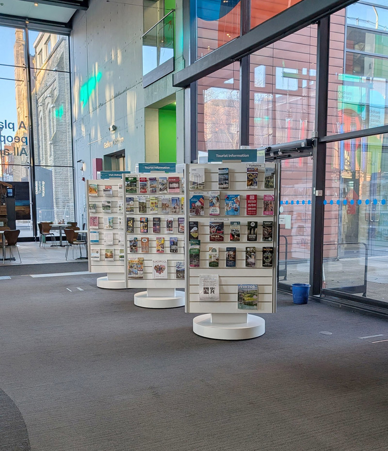 Three circular leaflet displays showcasing various brochures in a public library browsing area at Oldham Library