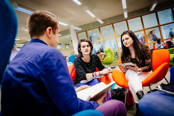 Bright orange tub chairs surround a collaborative work table where three students engage in discussion at Dublin City University