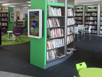 Purple lounge chairs and green study tables beside browser shelving in a digital offers area at Stafford Library