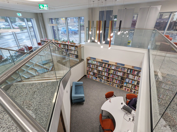 Curved glass railing and modern lighting above a study area with a round table and orange chairs at Stoke City Central Library