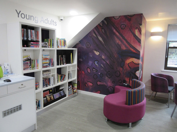 Colourful wall mural and purple lounge seating in a young adults' browsing area with shelving at Midsomer Norton Library