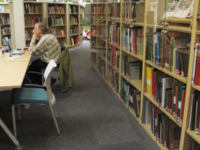 Study tables and a researcher in a quiet reading space beside glass-fronted shelving at Newark Local Studies