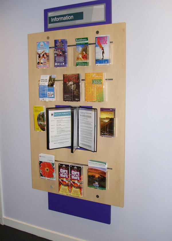 Wooden leaflet display board showcasing various brochures and guides in a public library browsing area at Morpeth Library