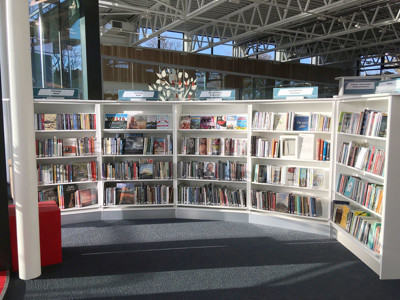Curved white shelving units filled with books in a public library browsing area at Conwy Culture Centre