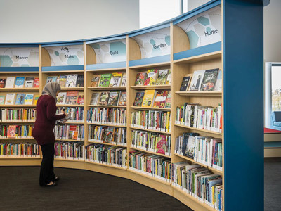 Curved shelving unit with themed book displays and a woman browsing titles in a public library browsing area at Sherwood Library