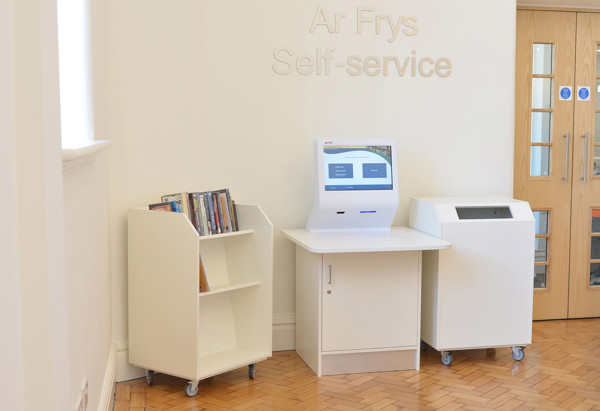 Self-service kiosk with a touchscreen and book return shelf beside a drop bin in a modern library space at Llandudno Library