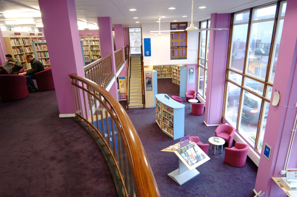 Curved wooden railing overlooking pink tub chairs and a browsing area with book displays at Ashford Library