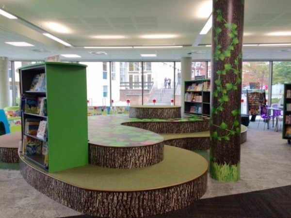Curved green seating areas with tree trunk designs and face-out book displays in a children's reading space at Stafford Library