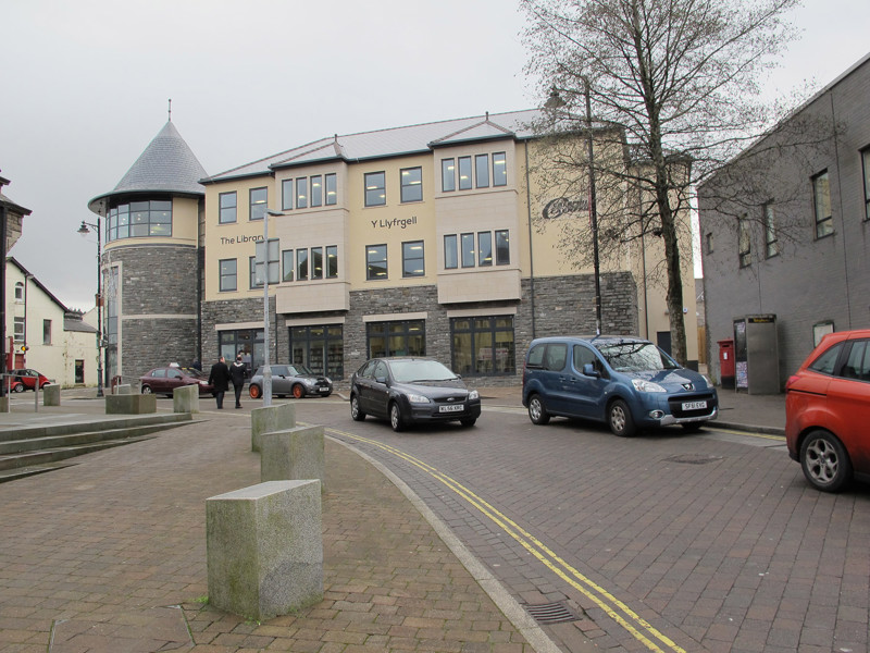 Modern stone and glass façade of a library building with a turret, surrounded by a paved area and parked cars at Caerphilly Library