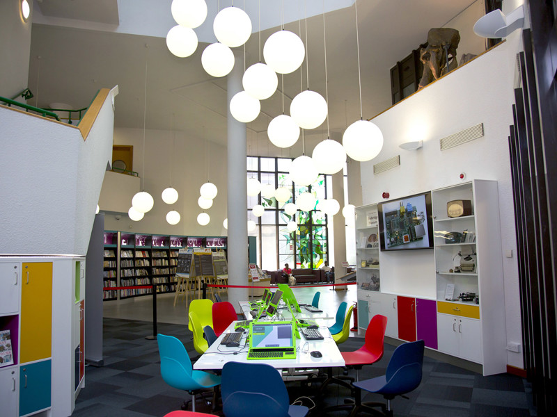 Brightly coloured chairs around a collaborative work table under large pendant lights in the makerspace at Redbridge Lab Central