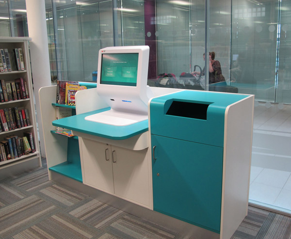 Turquoise self-service kiosk with a return shelf and drop bin beside browsing shelves at Orford Park Library