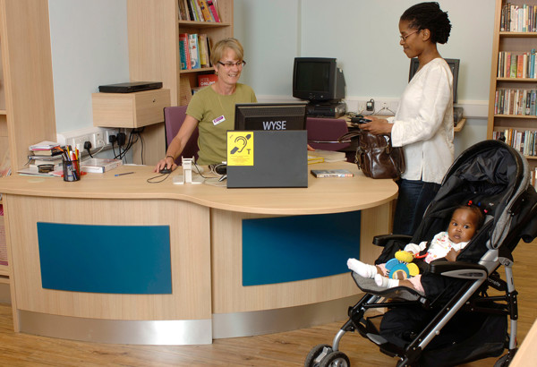 Curved wooden help desk with a staff member assisting a visitor and a child in a pushchair at Deanshanger Library