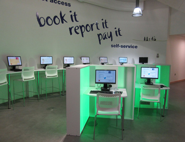 Self-service computer stations with illuminated green bases and white chairs in a public library browsing area at Southwater Customer Services
