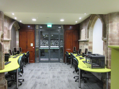 Curved green study tables with computer stations beside historic stone arches at Lichfield Library