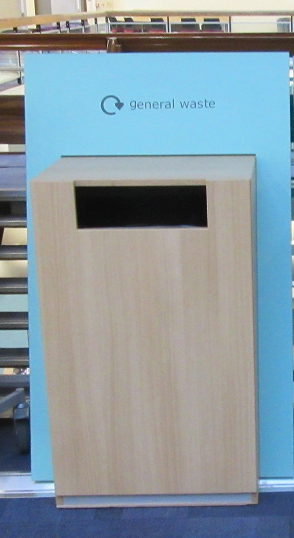 Wooden general waste bin with a sleek design and cut-out opening in a public library browsing area at County Library, Oxfordshire