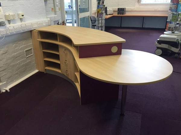 Curved wooden reception desk with shelving and a purple accent in the welcome zone at Broseley Library