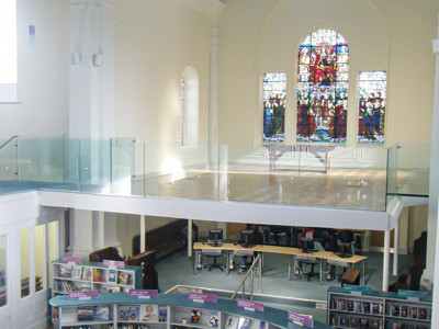 Stained glass windows illuminating the open-plan library floor with browsing shelves and study tables at St Aubyn Library Church