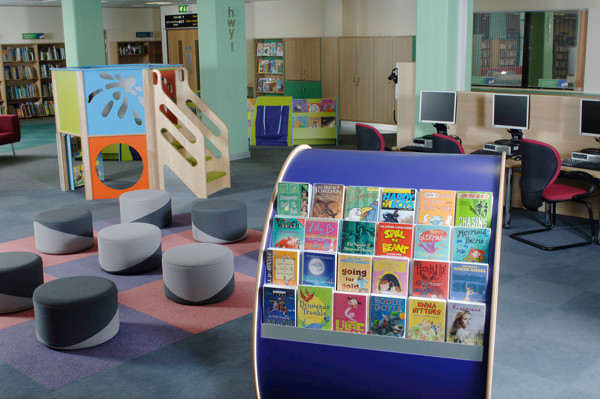 Face-out book displays featuring colourful children's titles beside soft grey seating in a playful reading area at Swansea Central Library