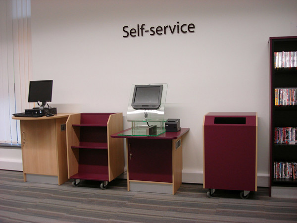 Self-service kiosk with a computer terminal, shelving unit, and drop bin in a library self-service area at Fitton Hill Library