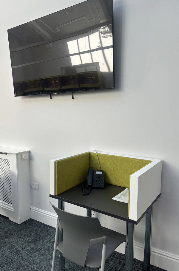 Grey study table with a green upholstered privacy screen and a phone, beneath a wall-mounted television at Royton Library