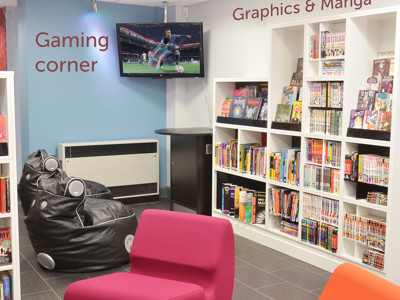 Bean bag seating and a wall-mounted screen in a gaming corner beside graphic novels at Gateshead Central Library