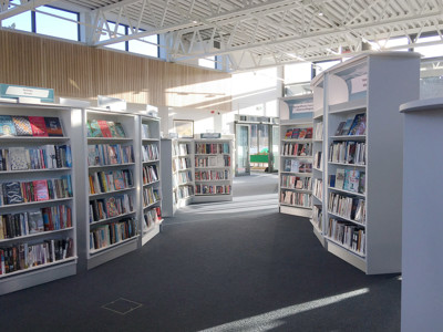 Curved browsing shelves filled with books creating a central pathway in a light-filled public library area at Conwy Culture Centre