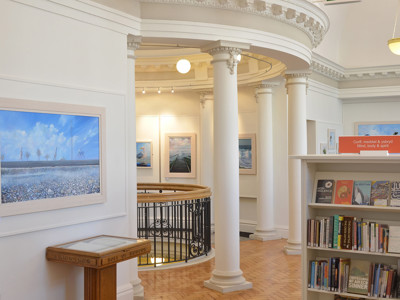 Curved wooden display table beside a bookcase and artwork in a light-filled reading area at Llandudno Library