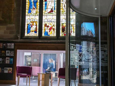 Stained glass windows illuminating a seating area with pink lounge chairs and a small table at Lichfield Library
