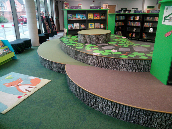 Curved seating areas with tree trunk designs and playful green flooring in a children's reading area at Stafford Library