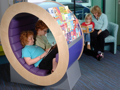 Two children reading inside a circular book display while a woman and another child read on a patterned sofa at Thame Library