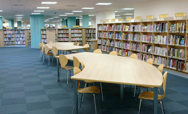 Curved wooden study tables surrounded by light wood chairs in a public library browsing area at Swansea Central Library