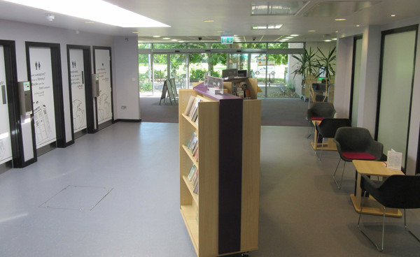 Open-plan library floor with a wooden book display and grey upholstered chairs at Wrexham Library