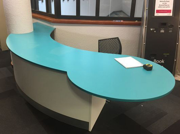 Curved turquoise reception desk with a black chair and book return unit at Redbridge Central Library