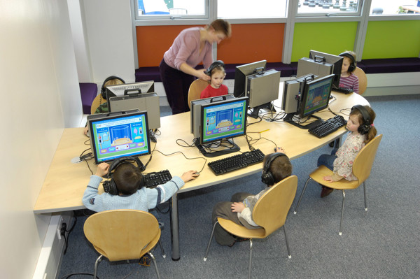 Children using computers with headphones at a study area featuring light-coloured tables and chairs at Milton Road Primary School