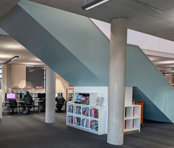 Curved wall and shelving unit beneath a staircase with study desks in the background at Oldham Library
