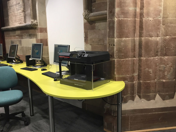 Bright yellow curved desk with a scanner and computer setup beside a stone wall at Lichfield Library