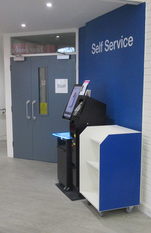 Self-service kiosk with a blue and white housing beside a staff door at Midsomer Norton Library