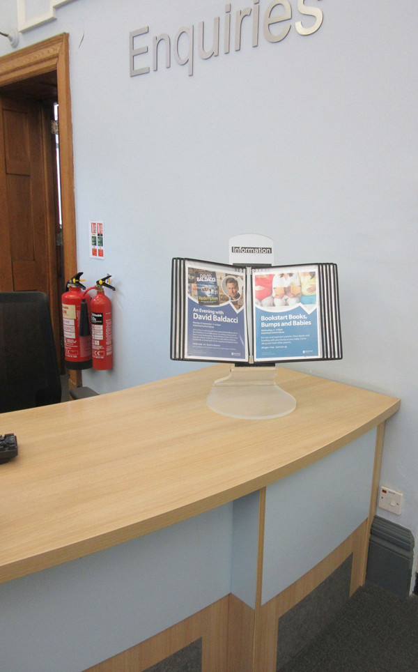 Curved wooden reception desk with a leaflet display stand featuring promotional materials at Gateshead Library