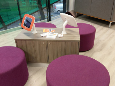 Purple upholstered stools surrounding a low wooden table with digital information screens at Newcastle-under-Lyme Library