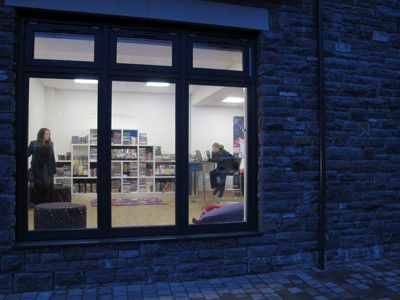 Two individuals engaged in activities at study tables beside browsing shelves in a well-lit space at Caerphilly Library