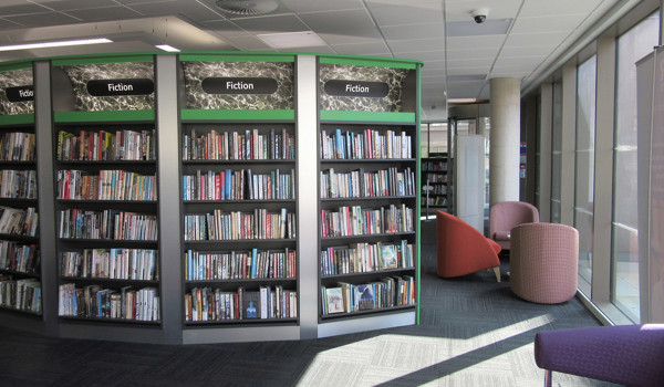 Curved shelving units filled with fiction titles beside upholstered seating areas at Stafford Library